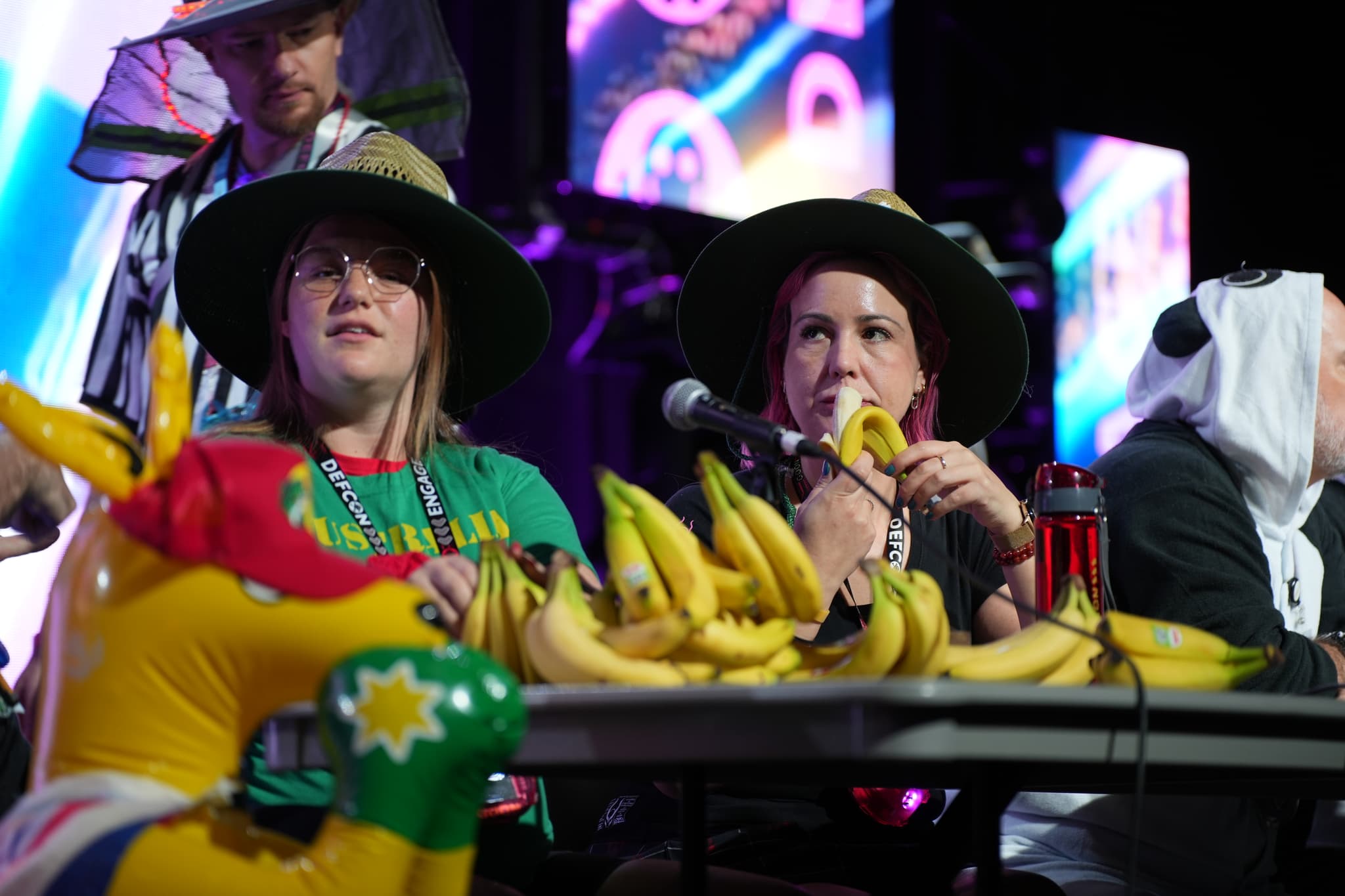 Two people wearing hats sit at a table covered with bananas, with one person holding a banana to their mouth. A person in the background is also wearing a hat, and there are colorful screens behind them