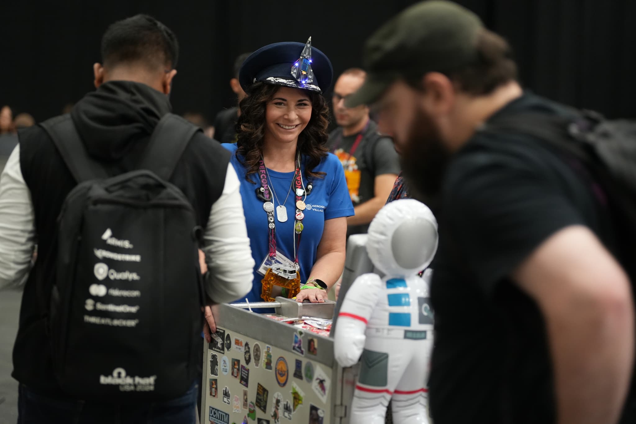 A woman in a blue shirt and a hat is smiling while interacting with people at an event. She is standing behind a table with a small astronaut figure on it. Several people are gathered around, some wearing backpacks