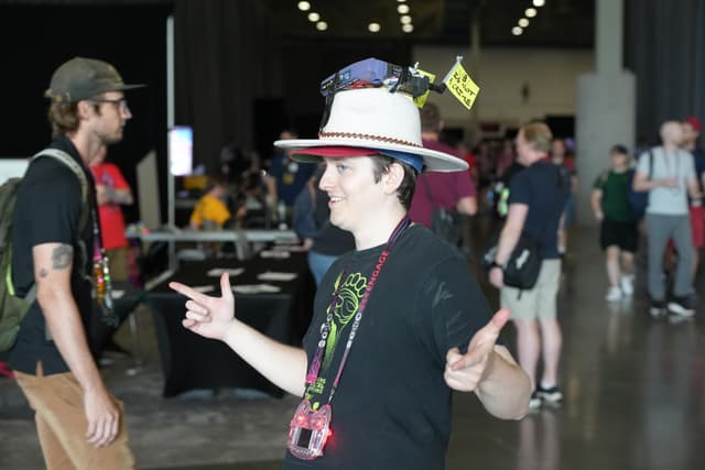 A person wearing a hat with multiple items attached to it, making finger guns gestures, in a crowded indoor event space