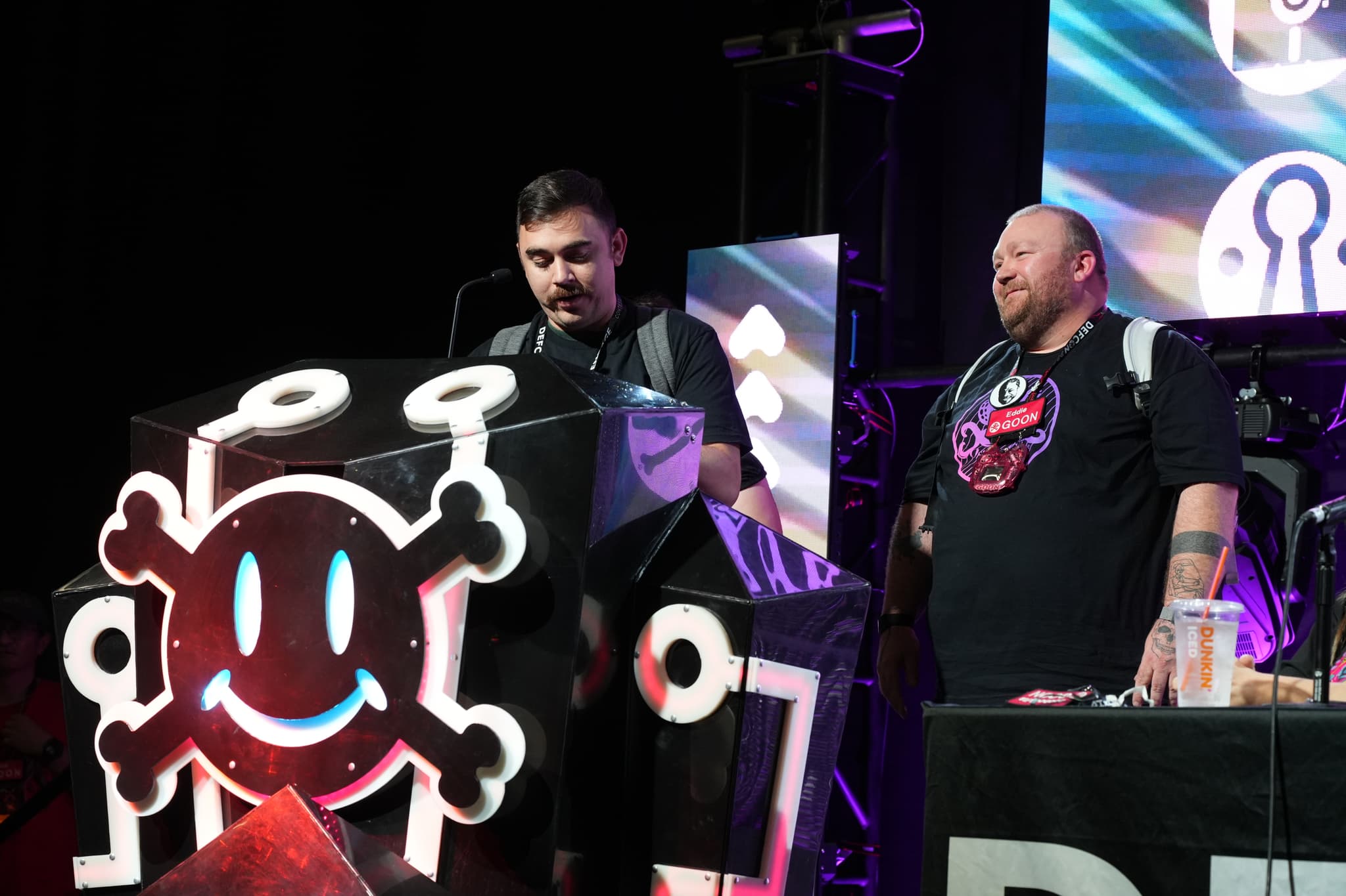 Two men standing on a stage, one speaking at a podium decorated with a smiling skull and crossbones symbol, and the other standing beside him. Both are wearing black shirts with similar designs. The background features colorful screens and lighting