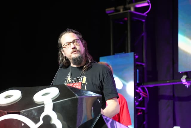 A man with long hair and glasses stands behind a podium, speaking at an event with stage lighting and screens in the background