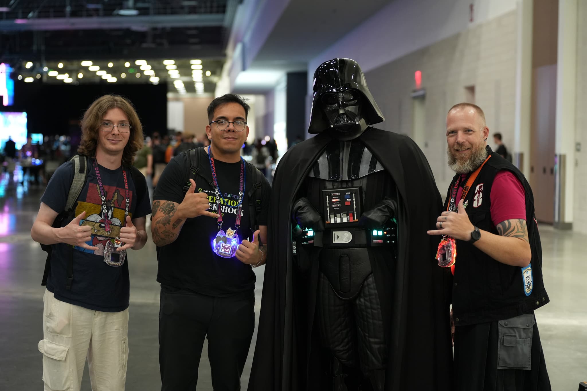 Four people standing together indoors, with one person dressed in a Darth Vader costume. The other three individuals are making hand gestures and smiling at the camera