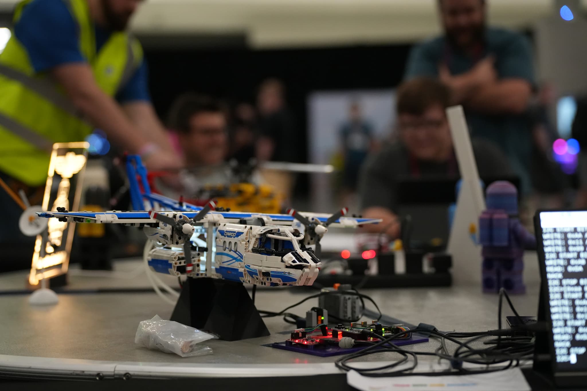 A table with a small helicopter model, various electronic components, and a computer screen displaying text, with people working in the background