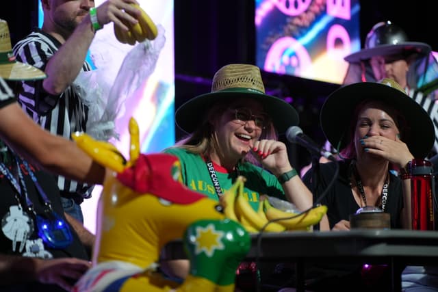 A group of people wearing hats and colorful attire, sitting at a table with inflatable toys and microphones, with a vibrant, illuminated background