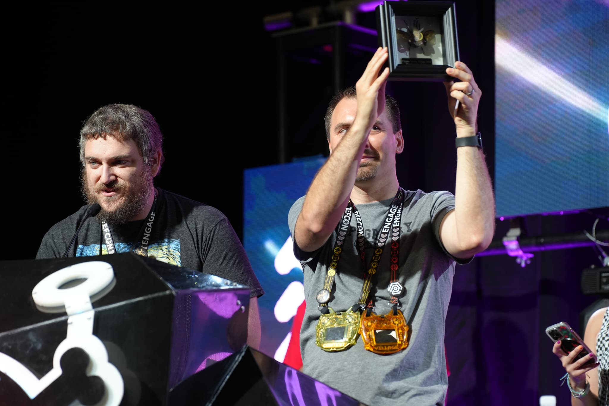 Two individuals on a stage, one holding up a trophy or award, with both wearing multiple medals around their necks