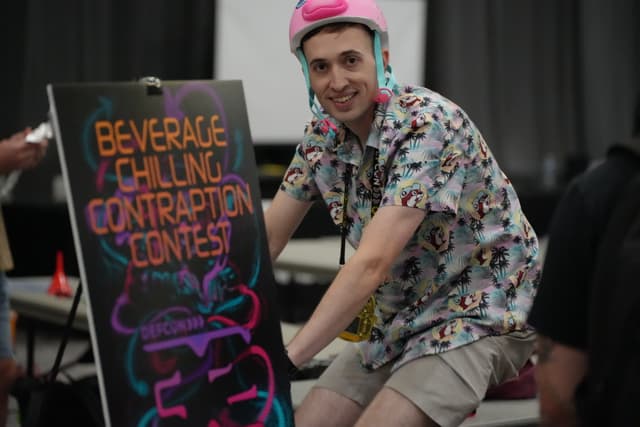 A person wearing a pink helmet and a patterned shirt is riding a stationary bike next to a colorful sign that reads Beverage Chilling Contraption Contest