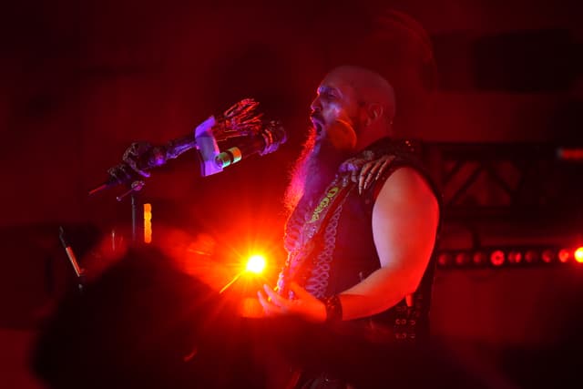 A musician with a long beard and bald head performs on stage under red lighting, holding a guitar and singing into a microphone