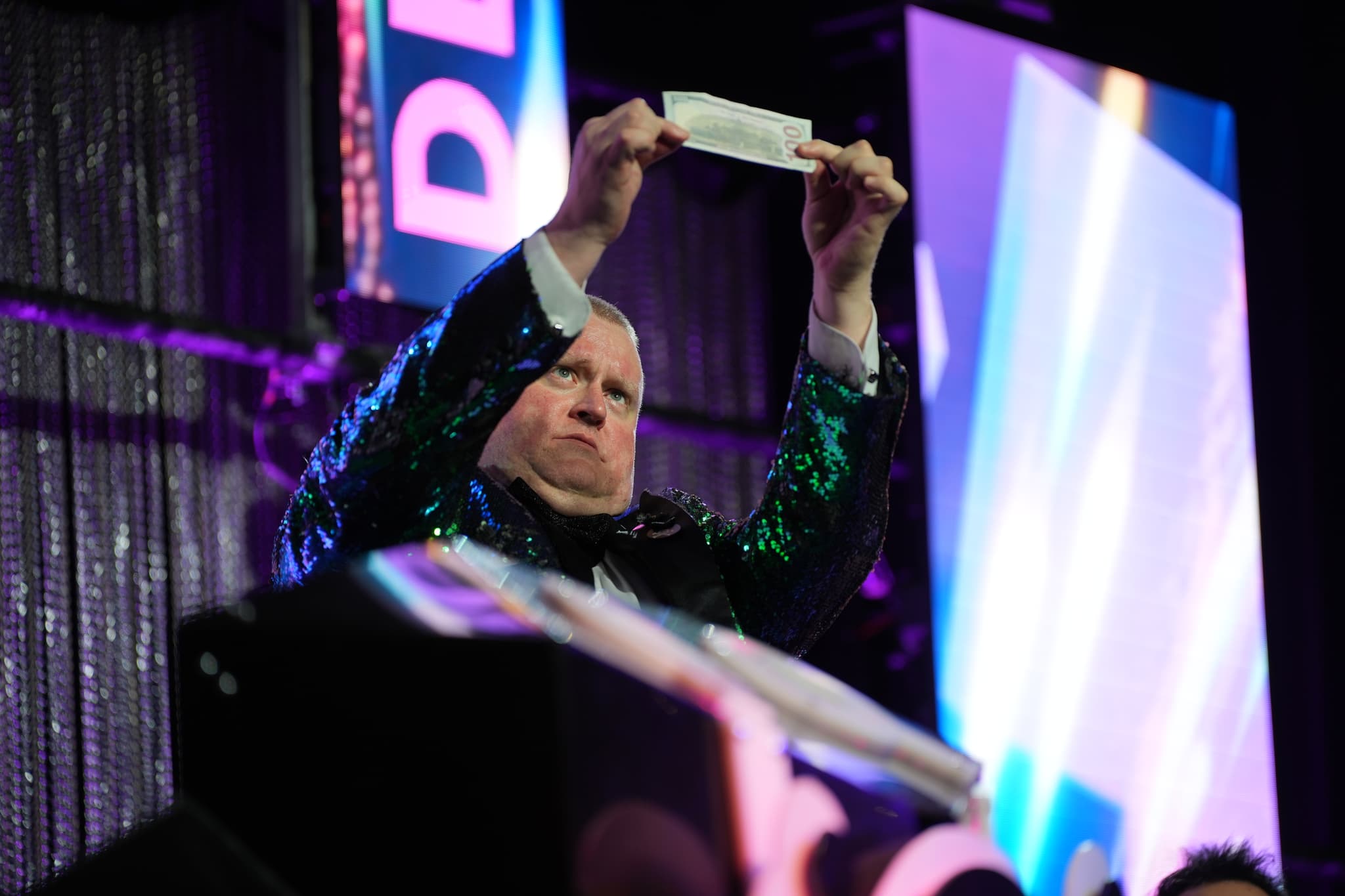 A person in a sparkly jacket holds up a piece of paper on a stage with colorful lighting and a backdrop