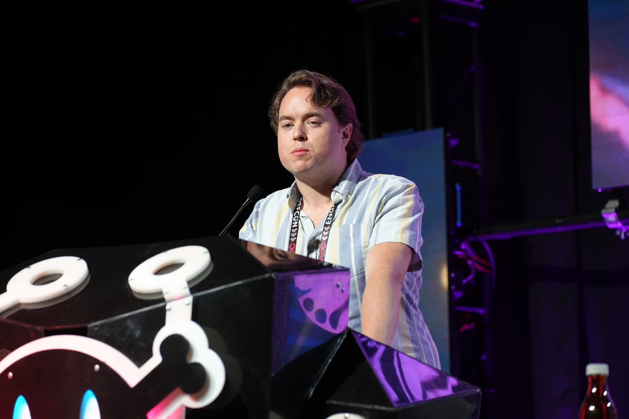 A person standing behind a podium with a microphone, wearing a light-colored shirt, and illuminated by stage lighting. The podium has a design featuring a stylized character with large eyes