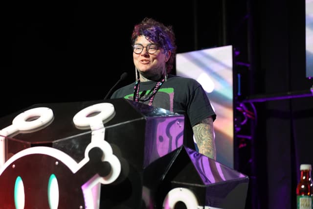 A person with glasses and colorful hair stands at a podium decorated with a skull and crossbones design, speaking into a microphone