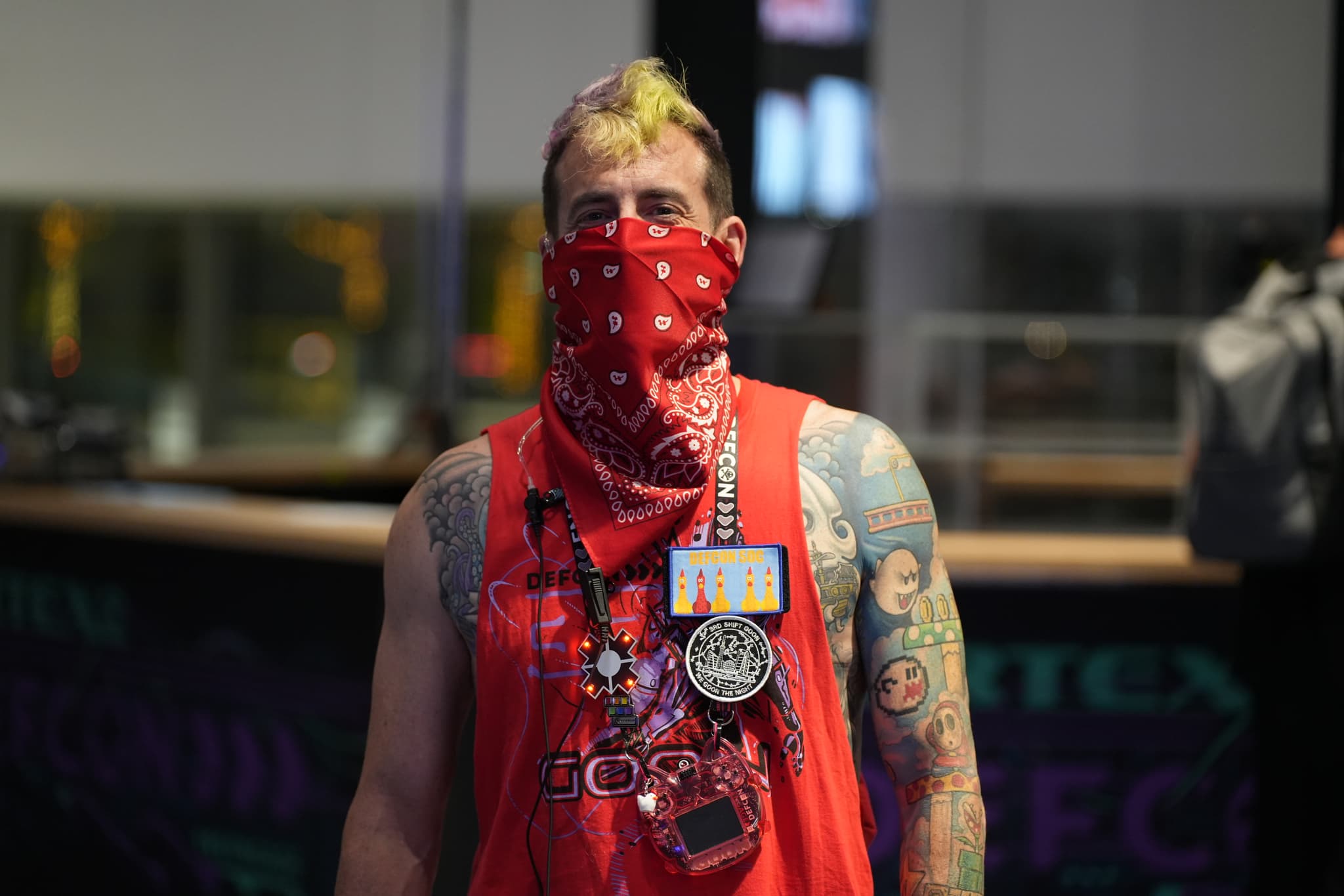 A person with a mohawk hairstyle and tattoos, wearing a red bandana over their face, a red tank top, and a lanyard with badges around their neck