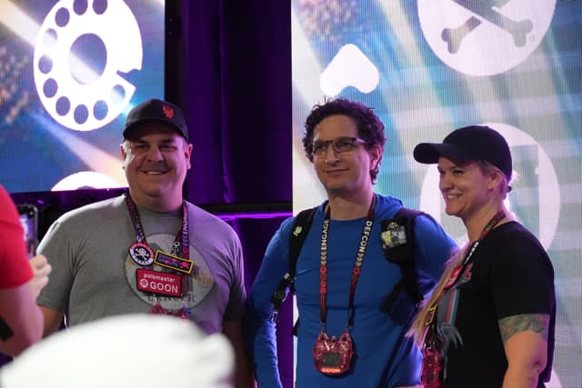 Three people standing together, smiling, and wearing lanyards with badges. The background features colorful, abstract designs and symbols