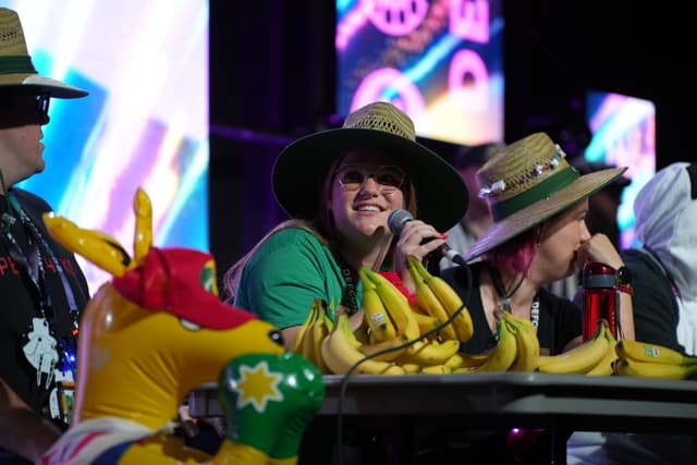 A group of people wearing hats and colorful outfits are sitting at a table with bananas and a kangaroo inflatable, with one person speaking into a microphone