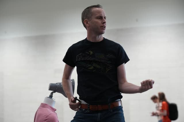 A person in a black t-shirt and jeans is striking a dynamic pose, possibly mimicking playing an air guitar. In the background, there is a mannequin with a pink shirt and a few blurred individuals