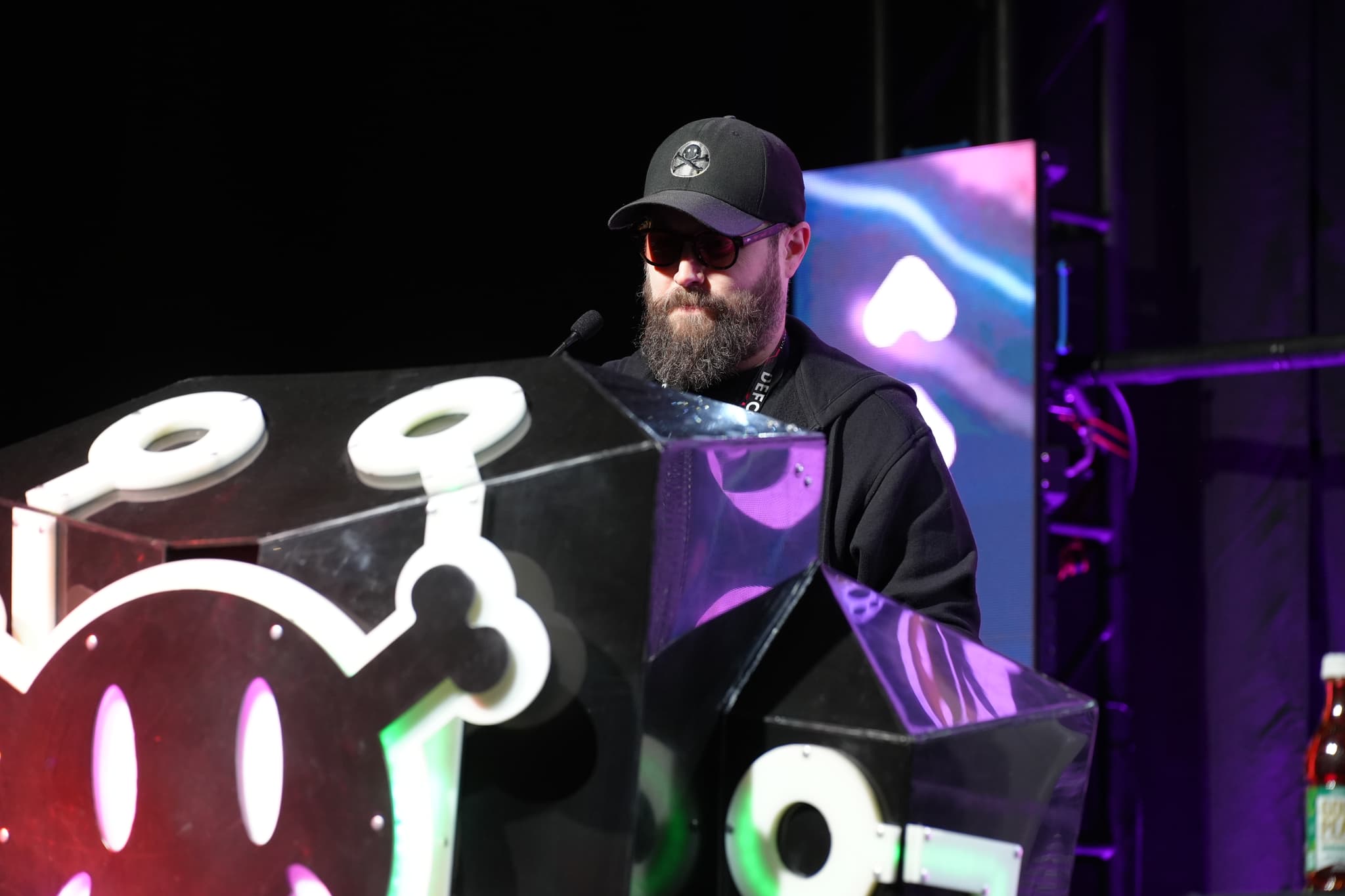 A person wearing a black cap and sunglasses stands behind a podium with a stylized design, possibly speaking or presenting at an event