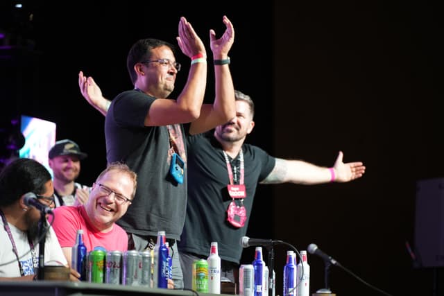 A group of people at a panel, with two individuals standing and gesturing energetically while others sit and smile. Various bottles and cans are placed on the table in front of them