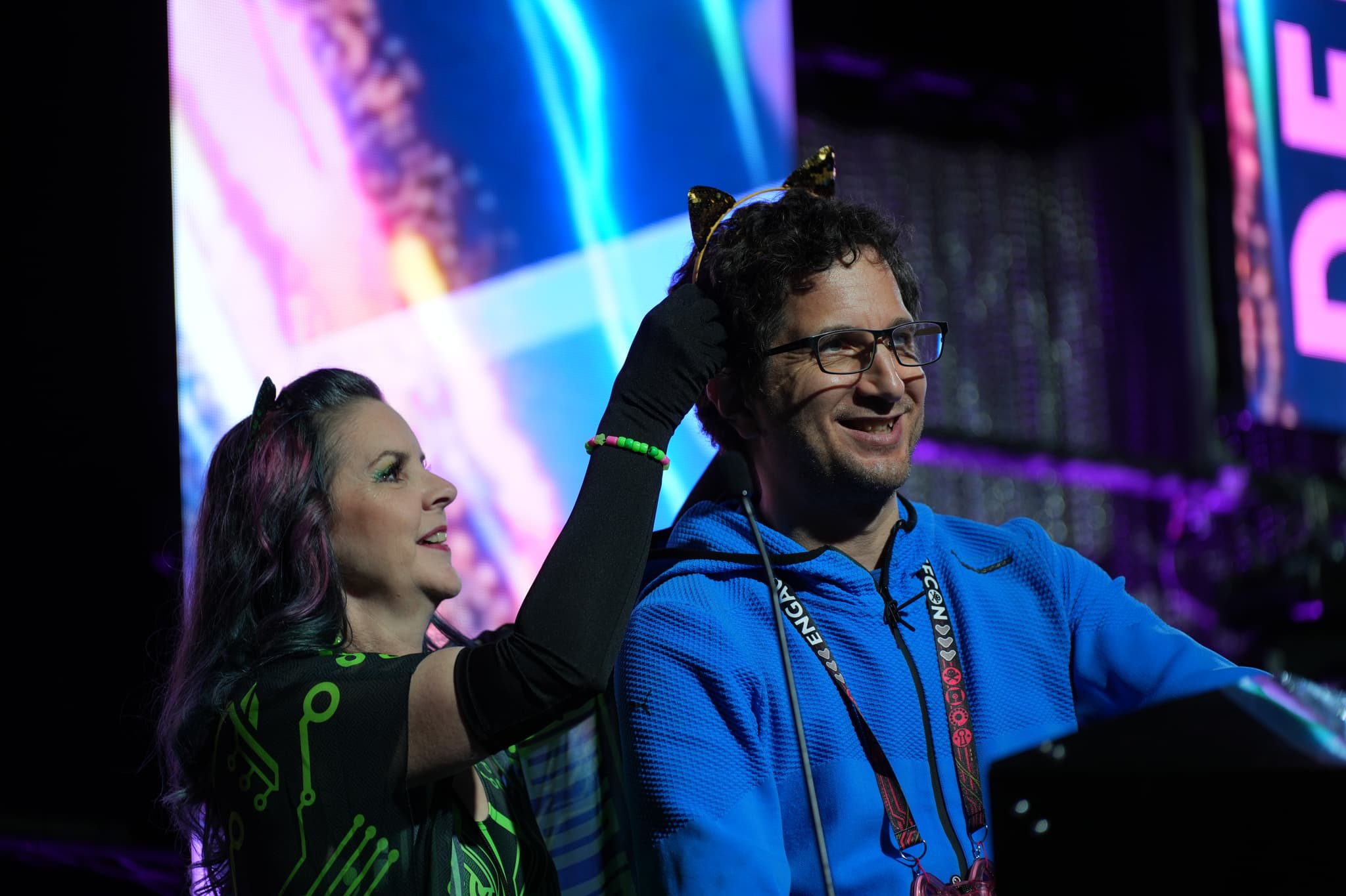 Two people are on stage, with one person placing cat ears on the other. The background features colorful, abstract lighting