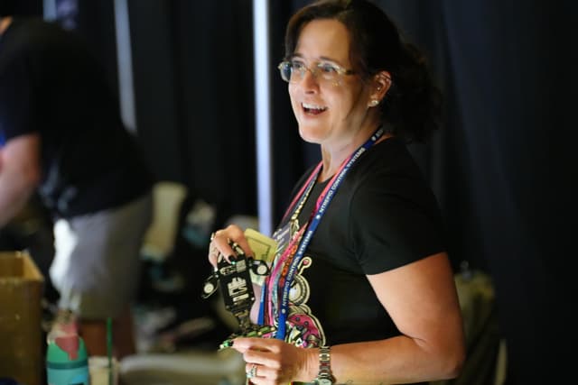 A woman with glasses and a black shirt is holding a camera and smiling, with a lanyard around her neck. She is indoors, and there is another person in the background