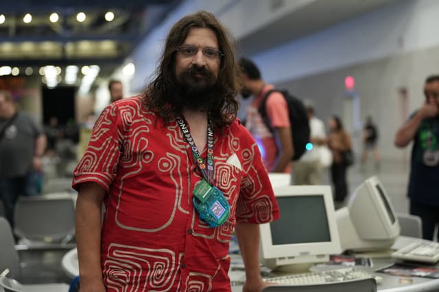 A man with long hair and a beard wearing a red patterned shirt stands in front of vintage computer equipment in a large indoor space with other people in the background