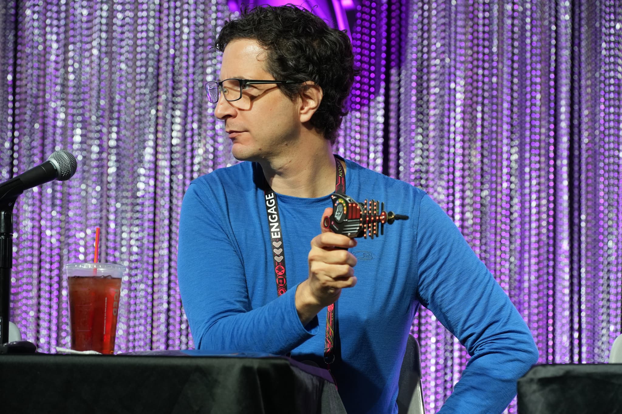 A person in a blue shirt holding a toy gun, sitting at a table with a microphone and a drink, against a backdrop of shimmering curtains