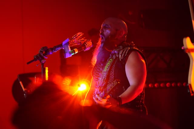 A musician with a beard and tattoos performs on stage under red lighting, playing a guitar and singing into a microphone