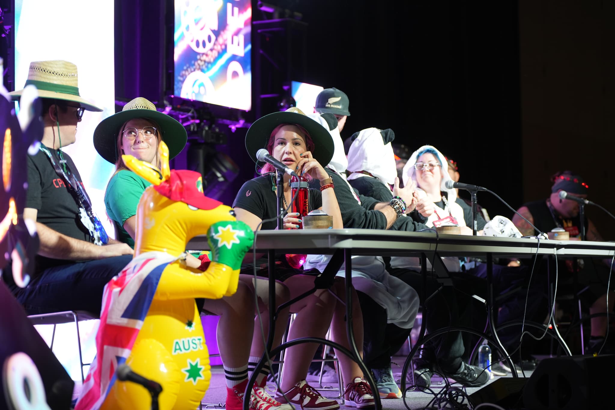 A group of people sitting at a table on a stage, wearing various hats and costumes, with microphones in front of them and a colorful inflatable toy in the foreground