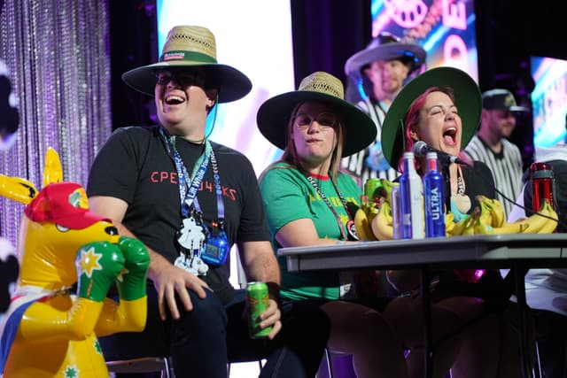 A group of people wearing hats and sitting at a table, with colorful decorations and a stuffed kangaroo in the foreground