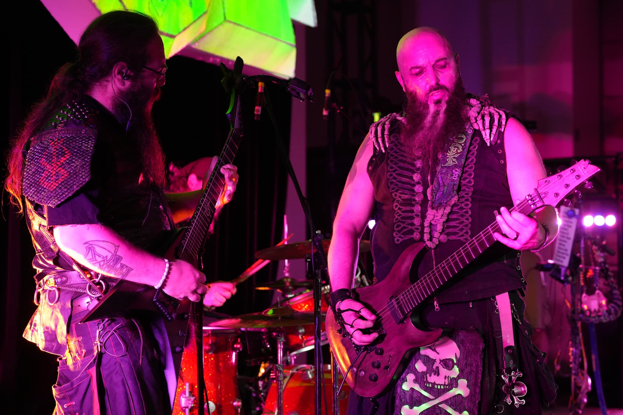 Two musicians performing on stage, both playing guitars, with vibrant lighting and a drum set in the background