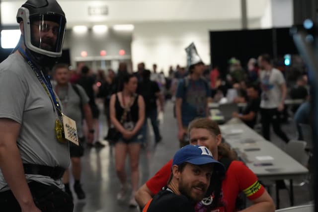 A crowded indoor event with people walking around and interacting. In the foreground, a person wearing a helmet and badge stands next to two seated individuals, one of whom is wearing a blue cap and red shirt. The background features tables and more attendees