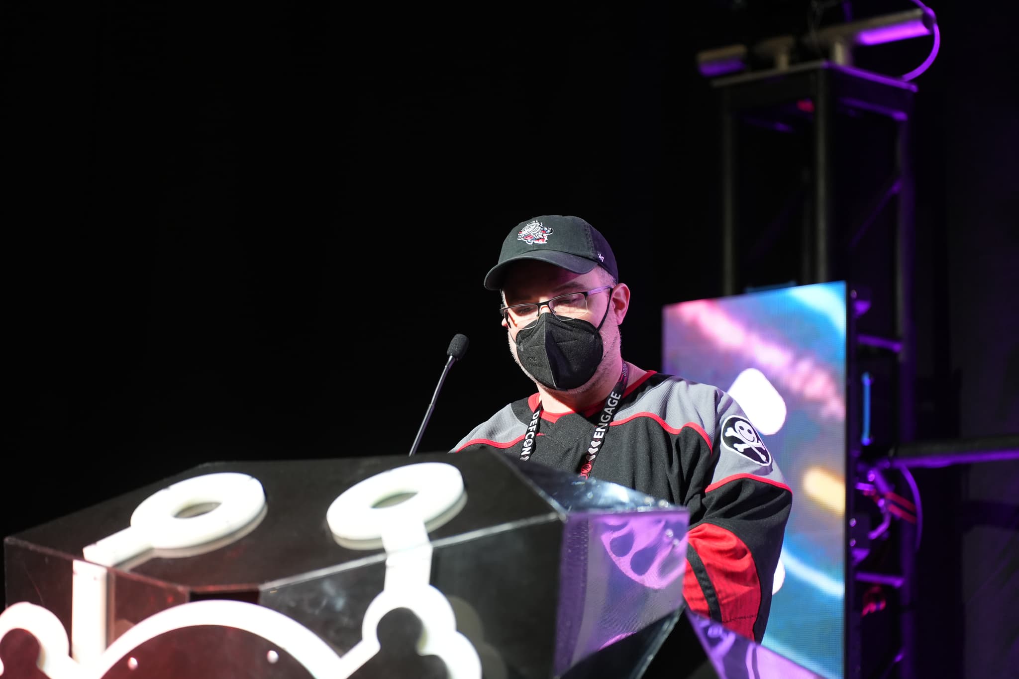 A person wearing a mask and cap stands behind a podium with a microphone, under stage lighting