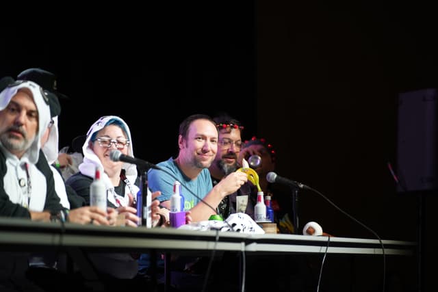 A group of people sitting at a table with microphones, some wearing costumes, and one person in the center smiling and holding a drink