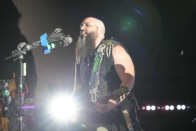 A musician with a bald head and a long beard performs on stage, illuminated by bright lights, wearing a studded vest and playing a guitar