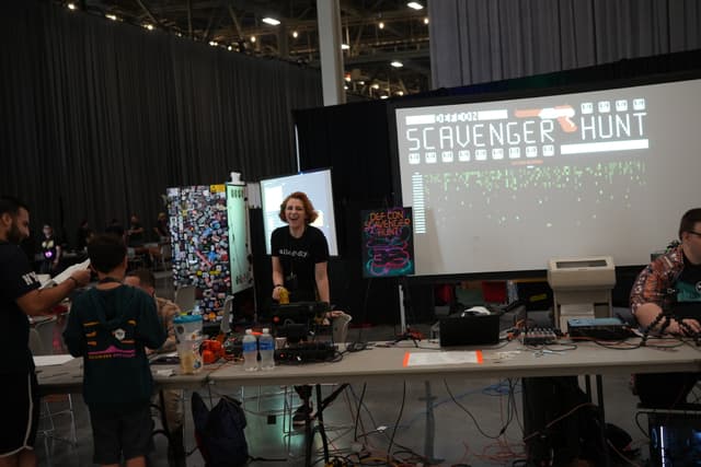 A group of people at a booth with a large screen displaying Scavenger Hunt in a convention or event setting. The booth has various equipment and items on the table, and the background includes a curtain and some colorful artwork