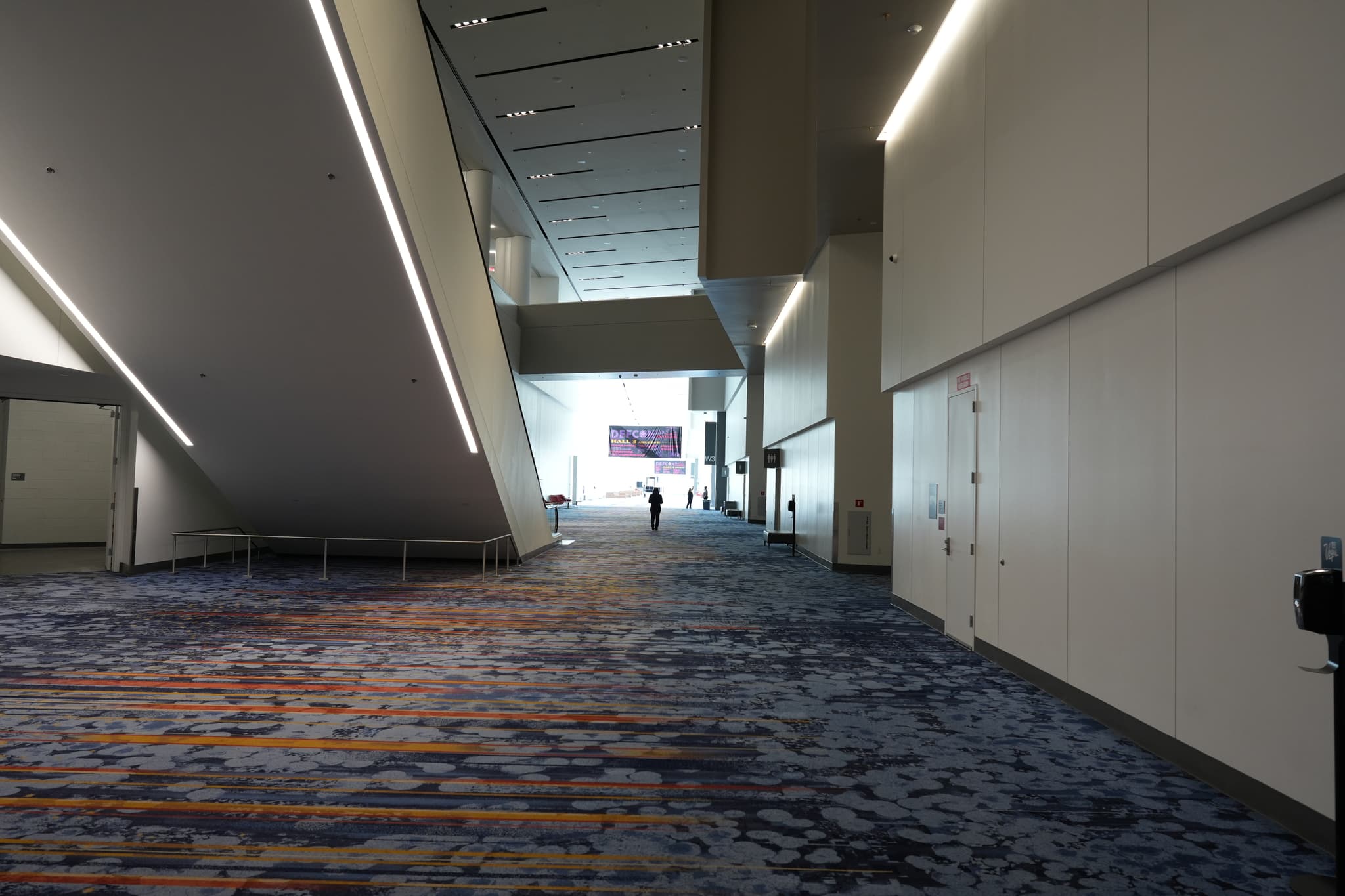 A spacious, modern hallway with high ceilings, patterned carpet, and minimalistic design, featuring a few people in the distance and illuminated by natural light from large windows