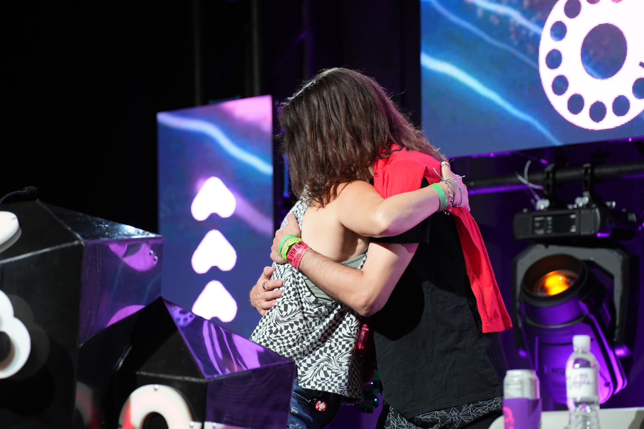 Two people hugging on a stage with a backdrop featuring heart symbols and circular designs, and a table with water bottles in the foreground