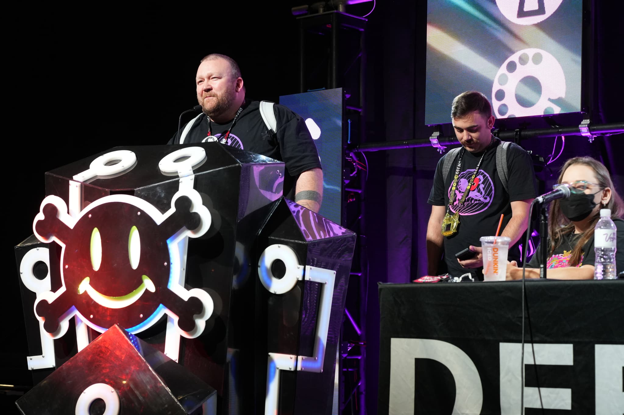 A group of people on a stage, with one person speaking at a podium decorated with a smiling skull and crossbones symbol. Two other individuals are seated at a table with the word DEF visible on a banner in front of them