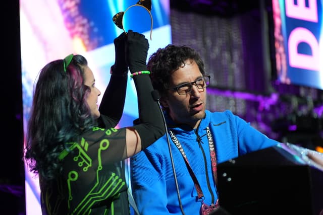 Two individuals on a stage, one holding a trophy above their head while the other looks on. The background features colorful lighting and a large screen