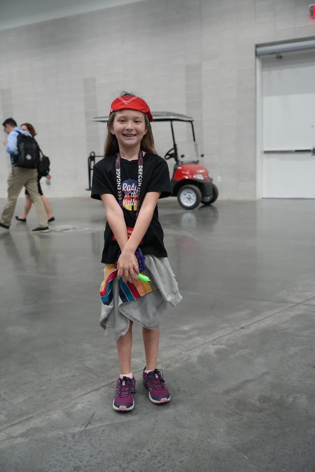 A young child wearing a red cap, black t-shirt, and gray skirt stands smiling in a large indoor space with a red golf cart and other people in the background