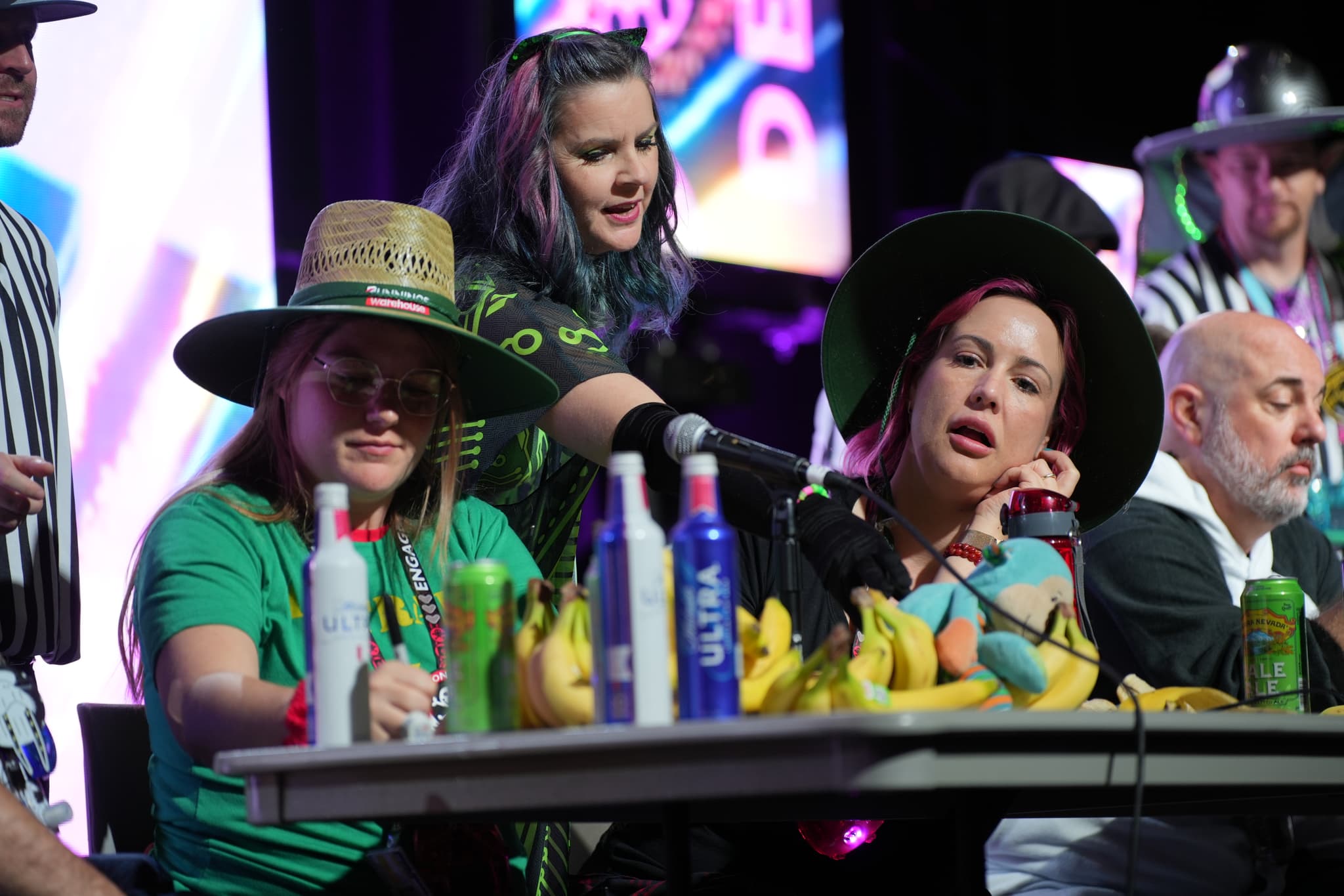 A group of people sitting at a table with bananas and various drinks, some wearing hats, and one person standing behind them with a microphone