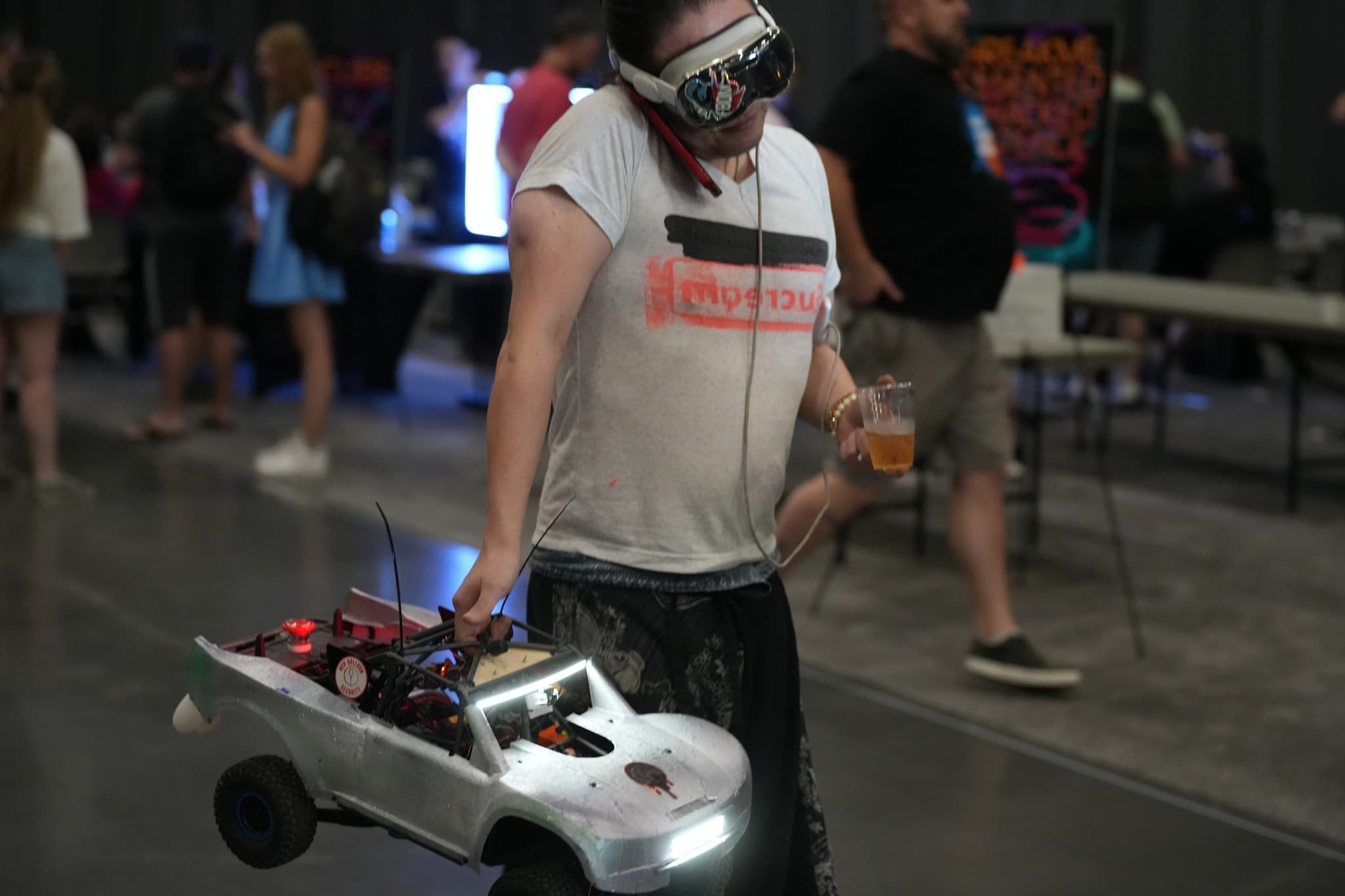 A person wearing virtual reality goggles and a white T-shirt is holding a remote-controlled car with electronic components on it, while standing in an indoor event space with other people in the background