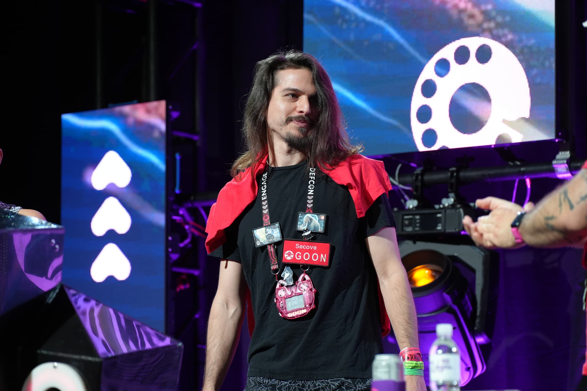 A man with long hair wearing a black t-shirt and a red jacket draped over his shoulders stands on a stage with colorful lights and screens in the background. He has a lanyard with badges around his neck and is looking to the side