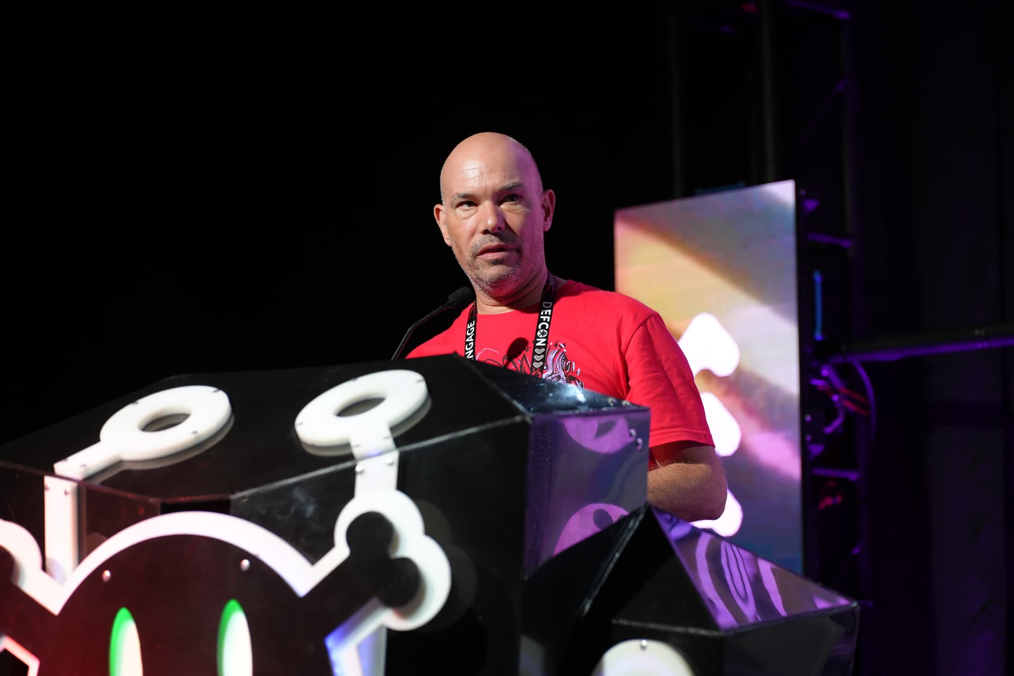 A person standing behind a podium decorated with a stylized skull and crossbones design, speaking into a microphone. The background features colorful lighting and abstract shapes