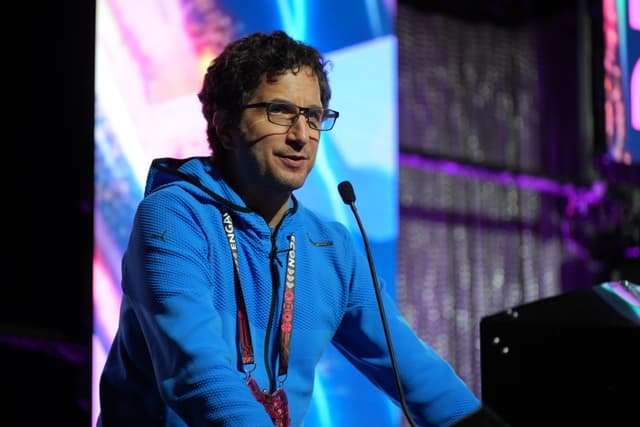 A person wearing glasses and a blue hoodie speaks into a microphone on a stage with colorful lighting in the background