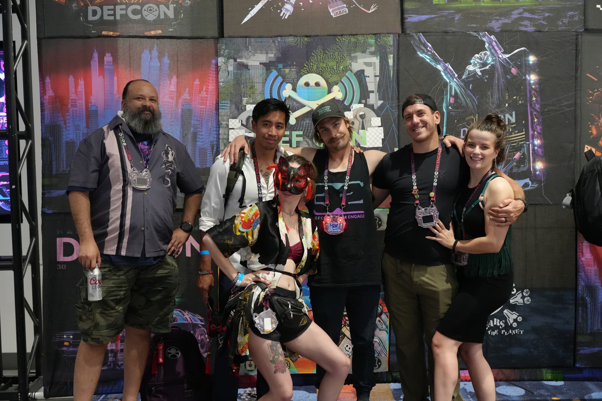 A group of six people posing together in front of a backdrop with various graphics and the word DEFCON visible. Some individuals are wearing lanyards and badges, and one person is dressed in a colorful outfit with a mask