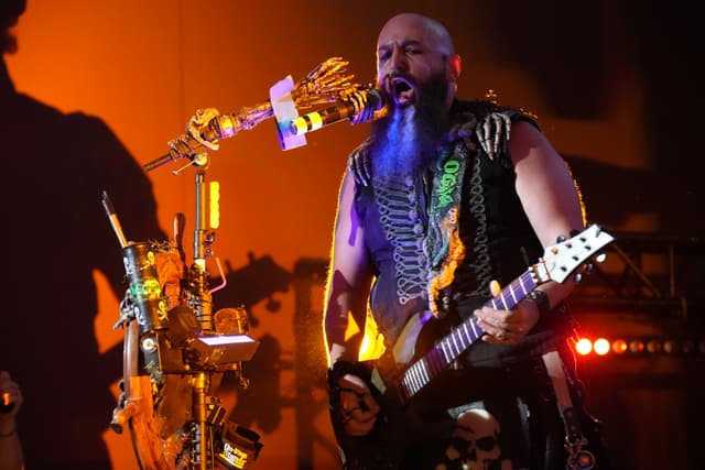 A musician with a blue beard and tattoos plays an electric guitar and sings into a microphone on stage, with colorful lighting and equipment in the background