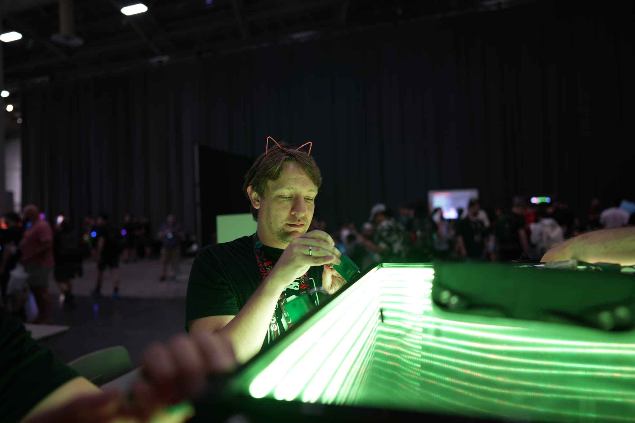 A person wearing cat ears is focused on an illuminated green object in a dimly lit room with other people in the background