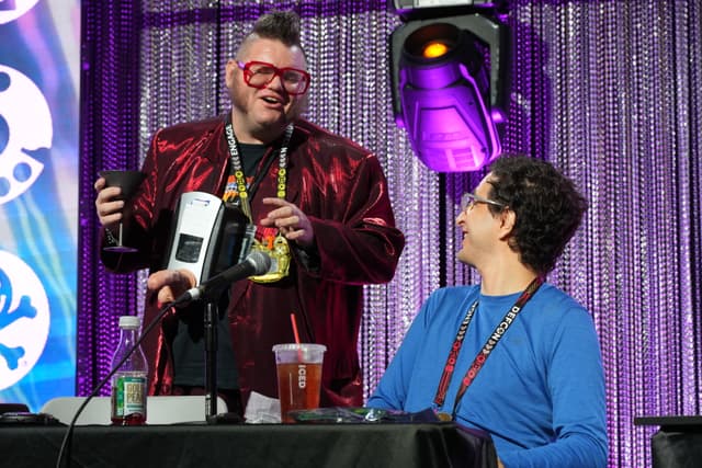 Two people at an event, one standing and holding a device while wearing a red jacket and large red glasses, and the other sitting and smiling, wearing a blue shirt. They are in front of a table with drinks and microphones, with a purple curtain and stage lights in the background