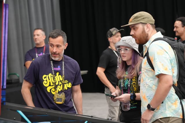 A group of people engaged in conversation at an event, with one person holding a controller and others wearing event badges and casual attire