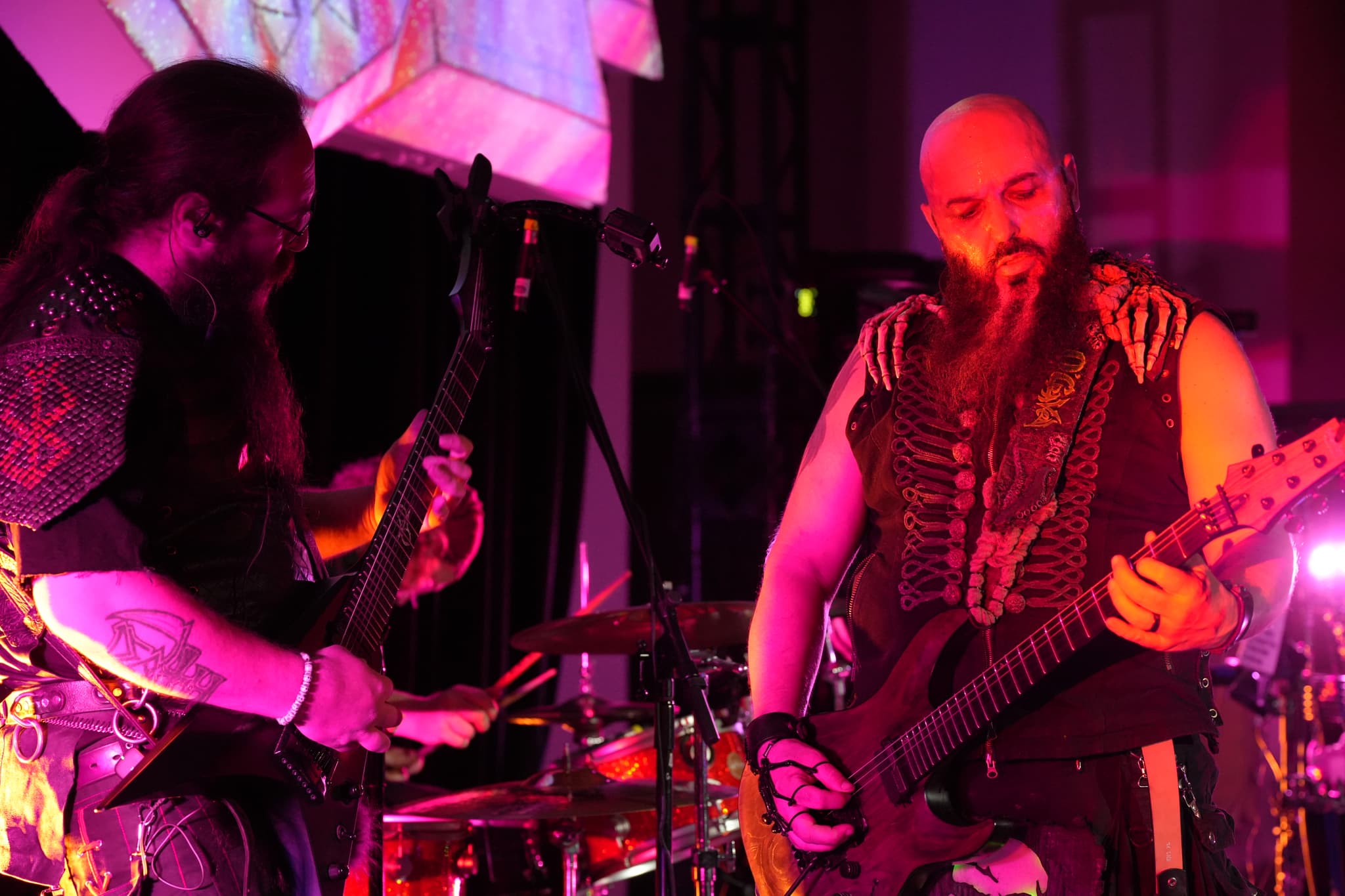 Two musicians performing on stage with electric guitars, illuminated by vibrant purple and pink lighting