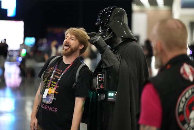 A man laughing and posing for a photo with someone dressed as Darth Vader, while another person looks on in the foreground